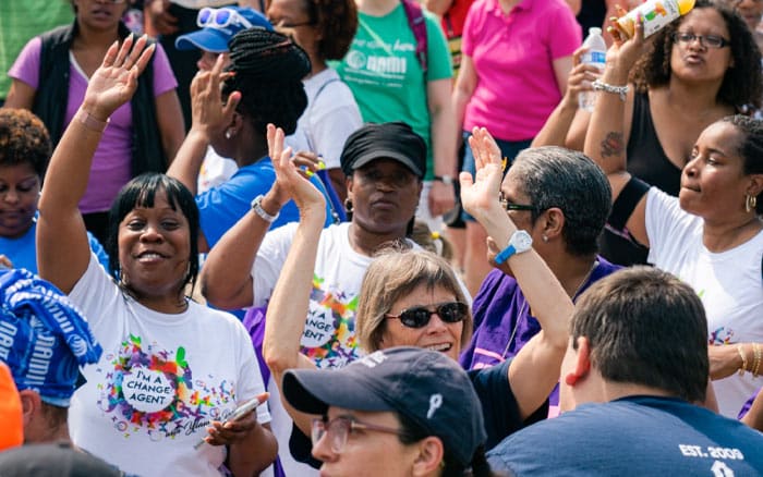 Two black ladies wearing a shirt saying "I'm a change agent" in the middle of a crowd looking at the camera with hands raised in the air